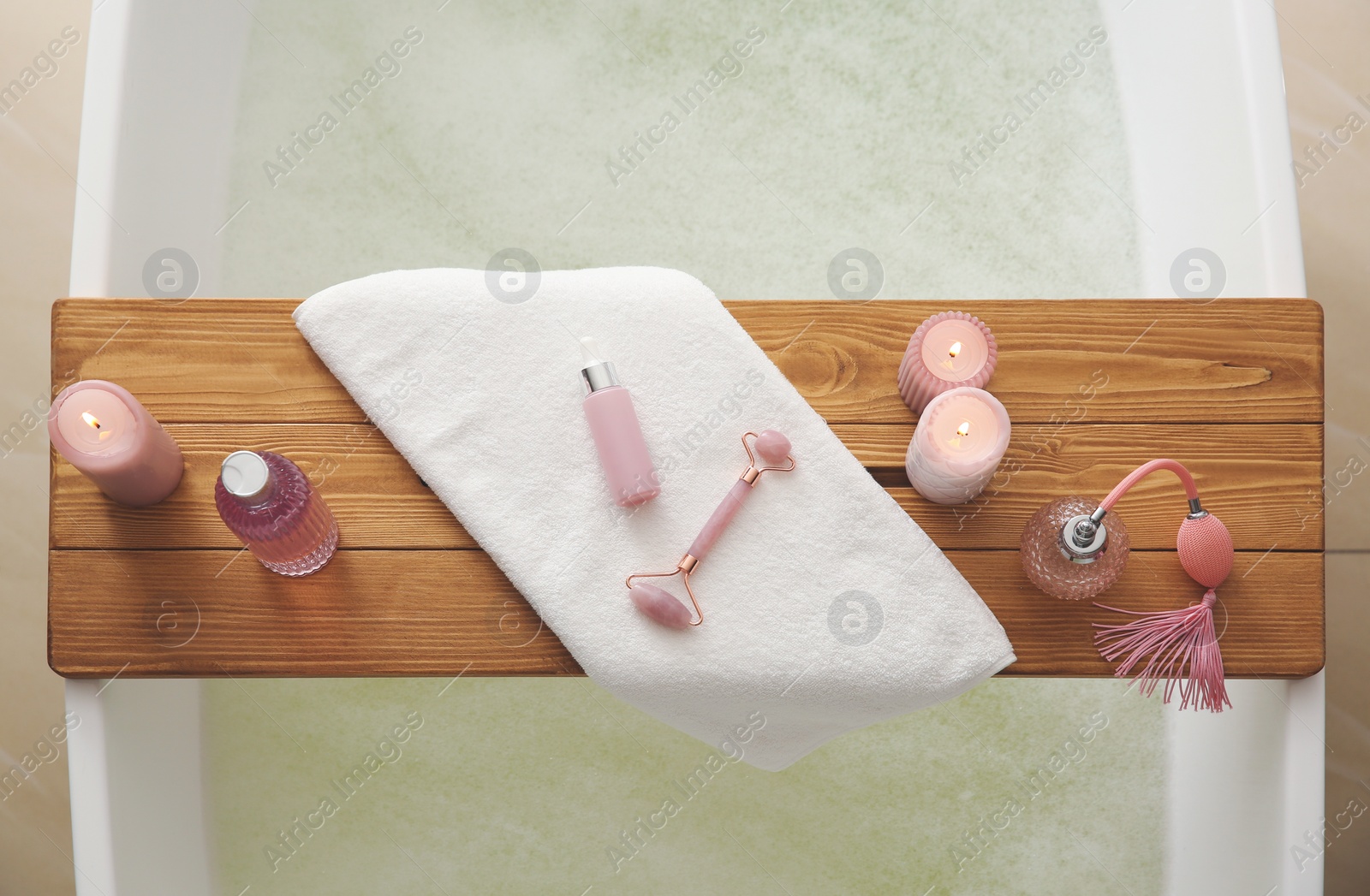 Photo of Wooden bath tray with face roller, cosmetic products and candles on tub indoors, top view
