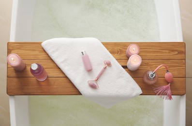 Wooden bath tray with face roller, cosmetic products and candles on tub indoors, top view