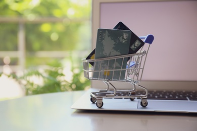 Photo of Internet shopping. Small cart with credit cards and laptop on table indoors, space for text
