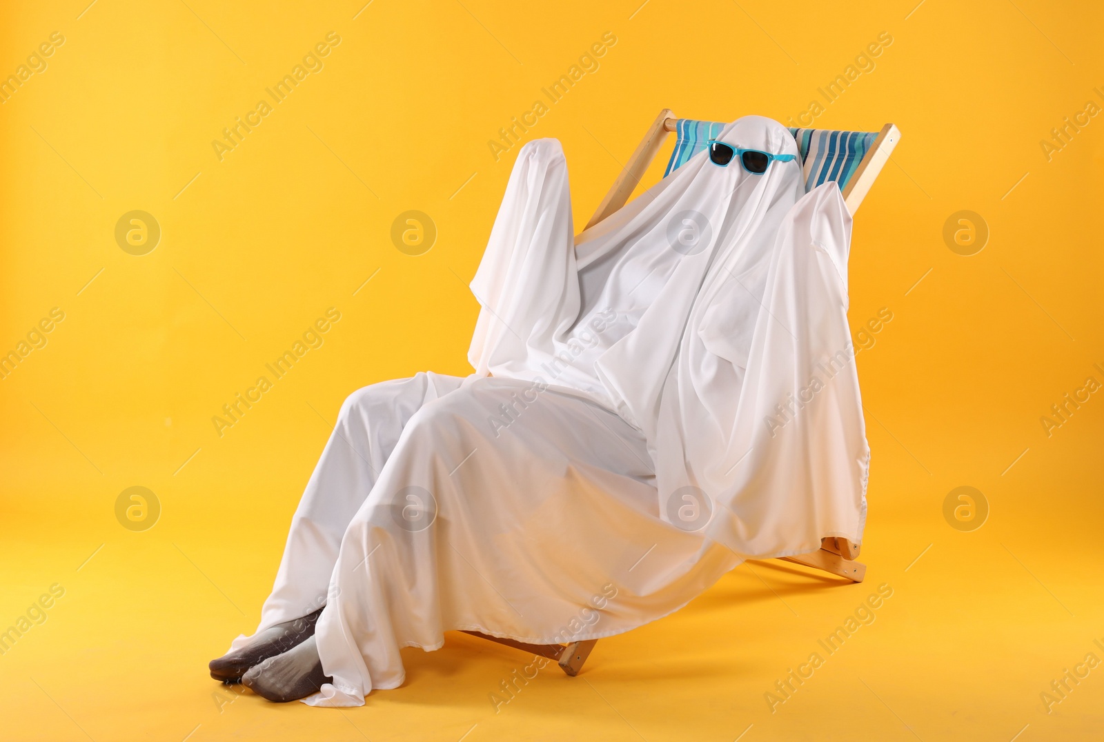 Photo of Person in ghost costume and sunglasses relaxing on deckchair against yellow background