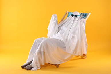Photo of Person in ghost costume and sunglasses relaxing on deckchair against yellow background