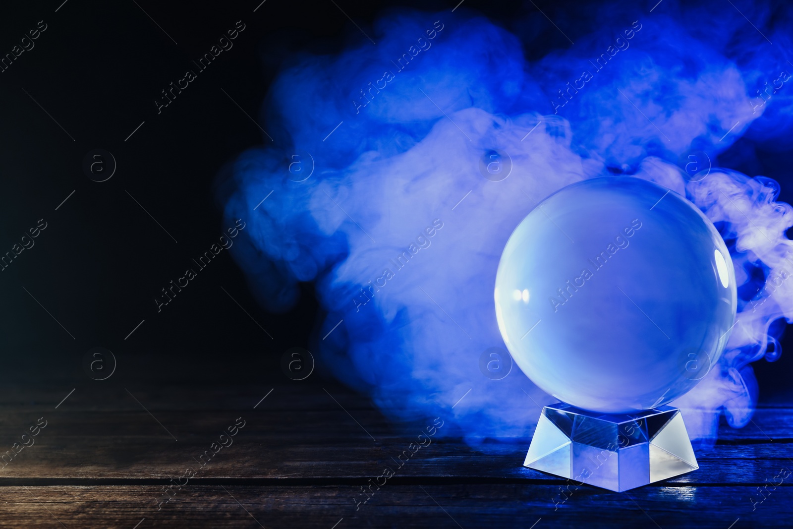 Photo of Magic crystal ball on wooden table and smoke against dark background. Making predictions