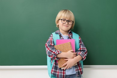 Photo of Happy little school child with notebooks near chalkboard. Space for text
