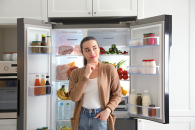 Photo of Thoughtful young woman near open refrigerator in kitchen