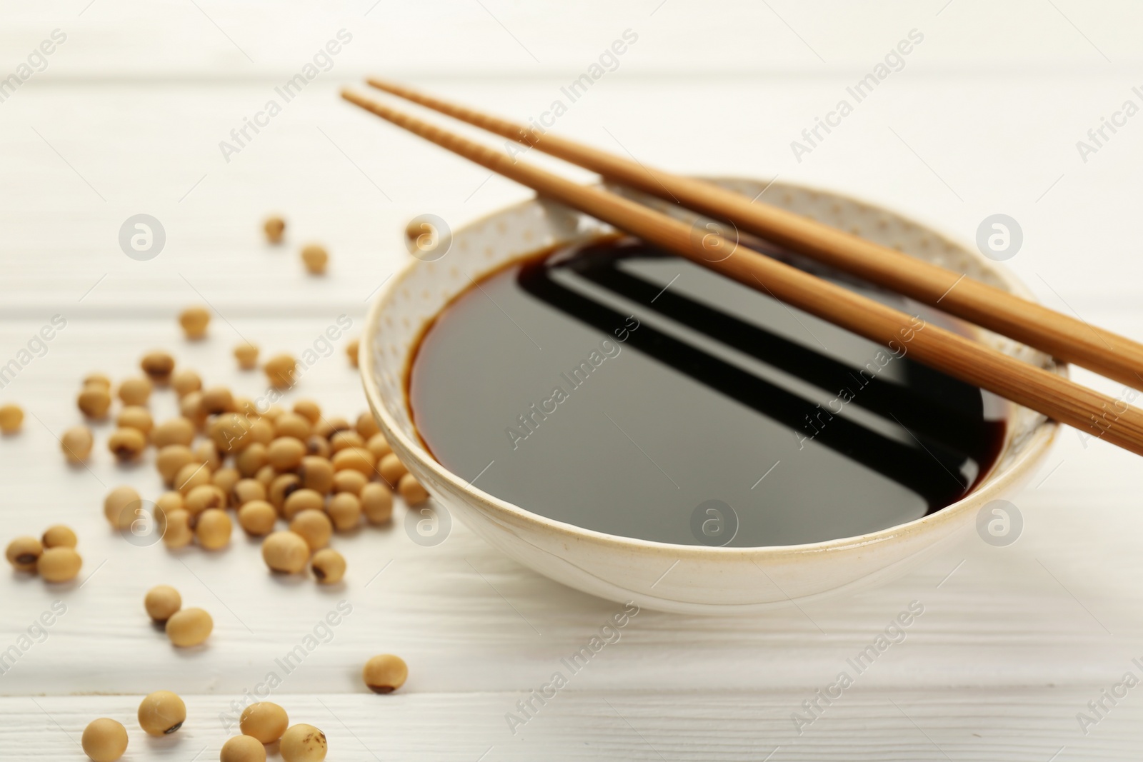 Photo of Tasty soy sauce in bowl, chopsticks and soybeans on white wooden table, closeup