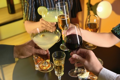 Photo of People with different alcohol drinks clinking glasses indoors, closeup