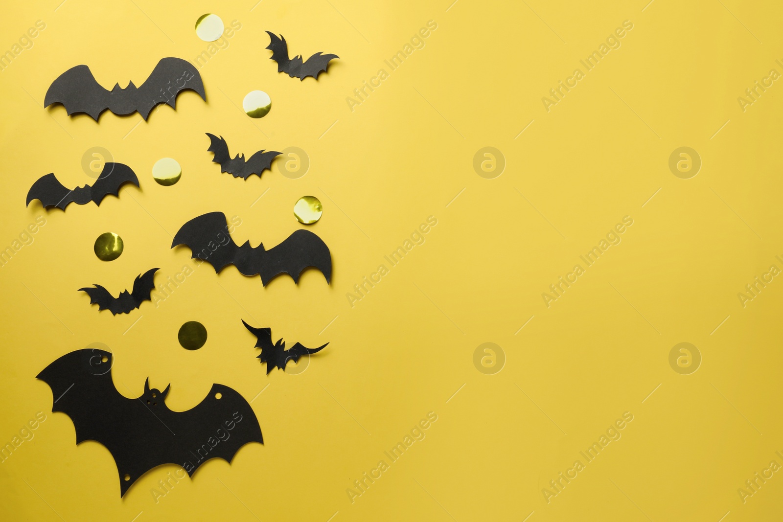 Photo of Flat lay composition with paper bats and golden confetti on pale yellow background, space for text. Halloween celebration