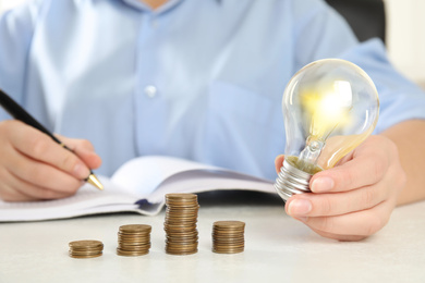 Photo of Woman with light bulb, notebook and coins at white table, closeup. Power saving
