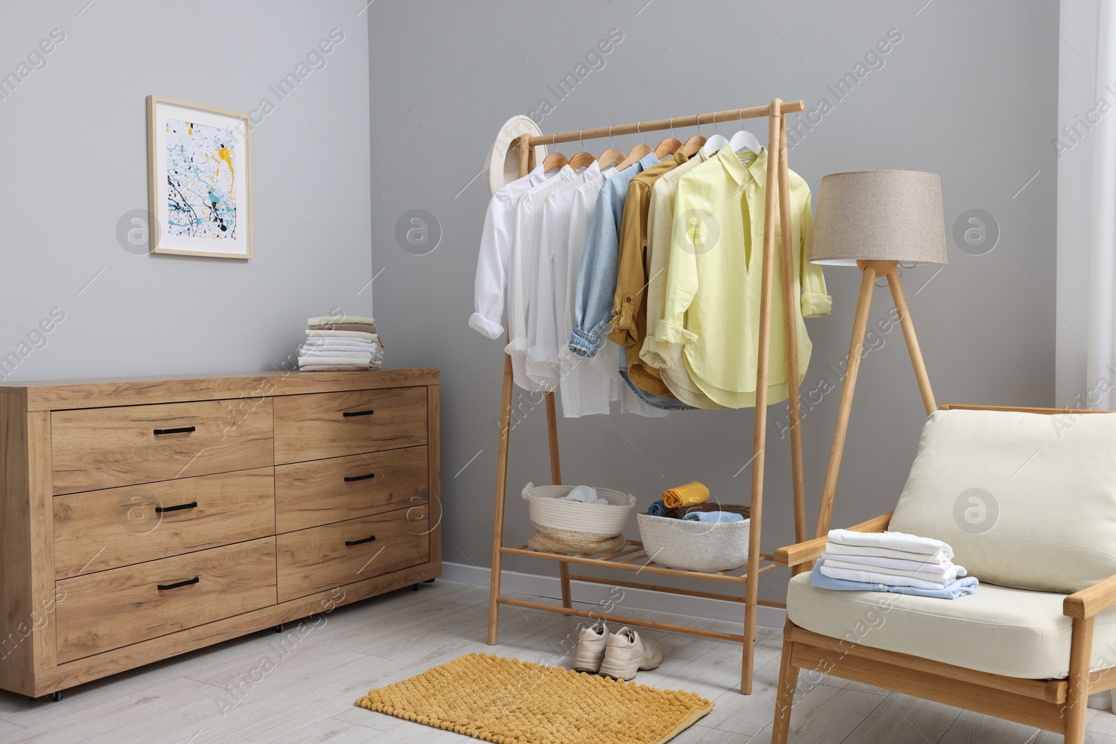 Photo of Wardrobe organization. Rack with different stylish clothes, chest of drawers, armchair and lamp near grey wall indoors