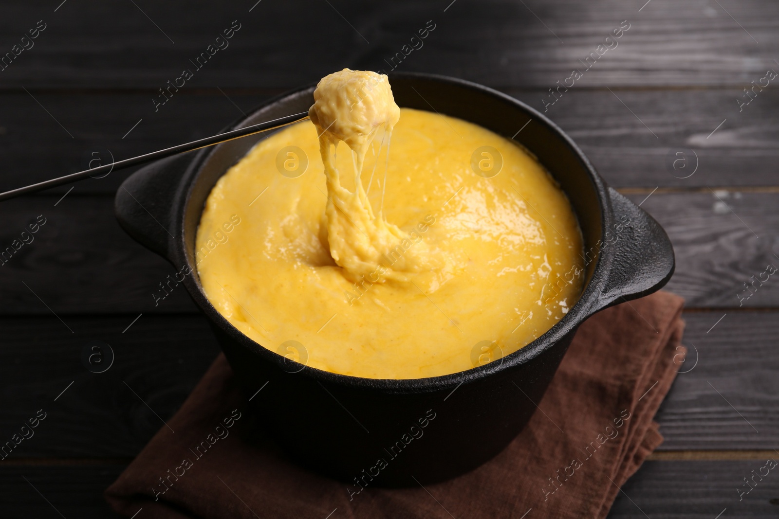 Photo of Dipping piece of ham into fondue pot with melted cheese on black wooden table, closeup