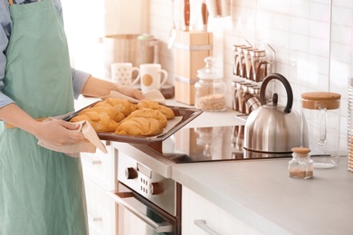 Woman holding baking tray with delicious croissants in kitchen, closeup