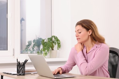 Woman suffering from pain in shoulder while using laptop at wooden table indoors. Arthritis symptoms