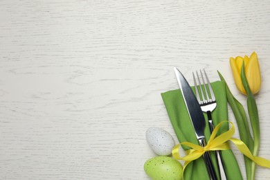 Photo of Cutlery set, painted eggs and beautiful flower on white wooden table, flat lay with space for text. Easter celebration