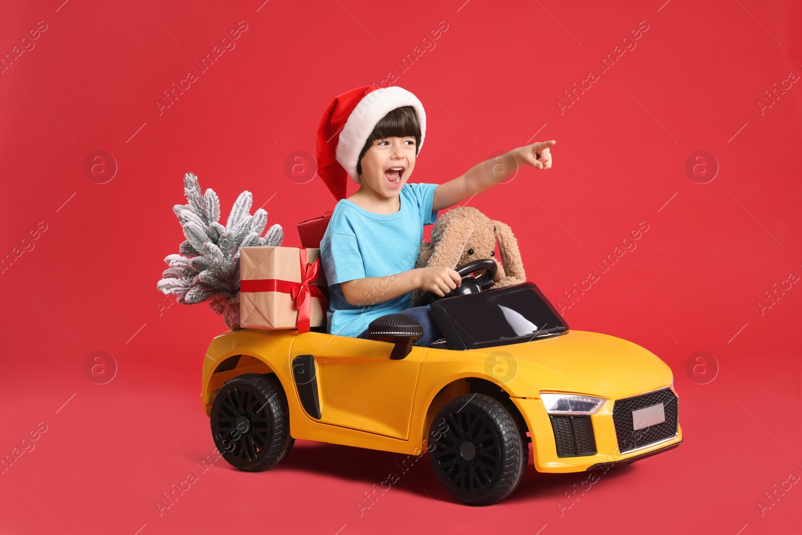Photo of Cute little boy in Santa hat with Christmas tree, bunny toy and gift box driving children's car on red background