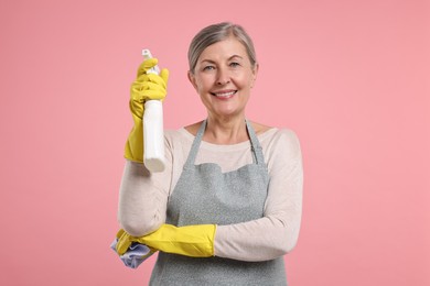 Happy housewife with spray bottle and rag on pink background