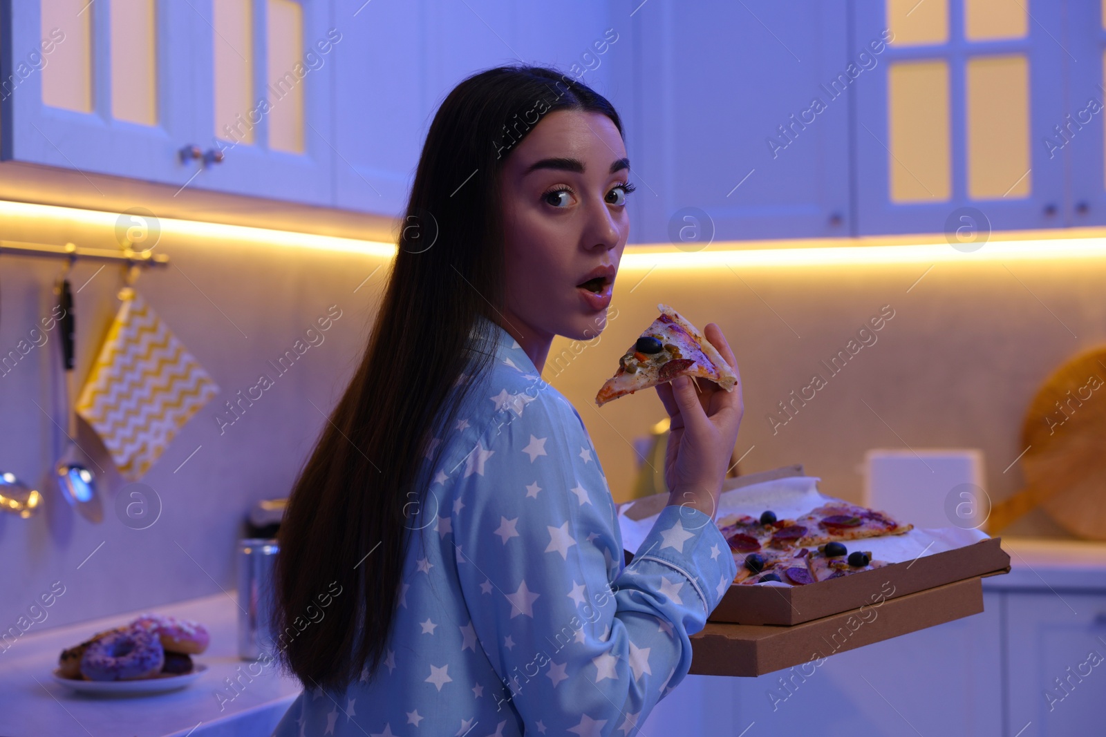 Photo of Young woman eating pizza in kitchen at night. Bad habit