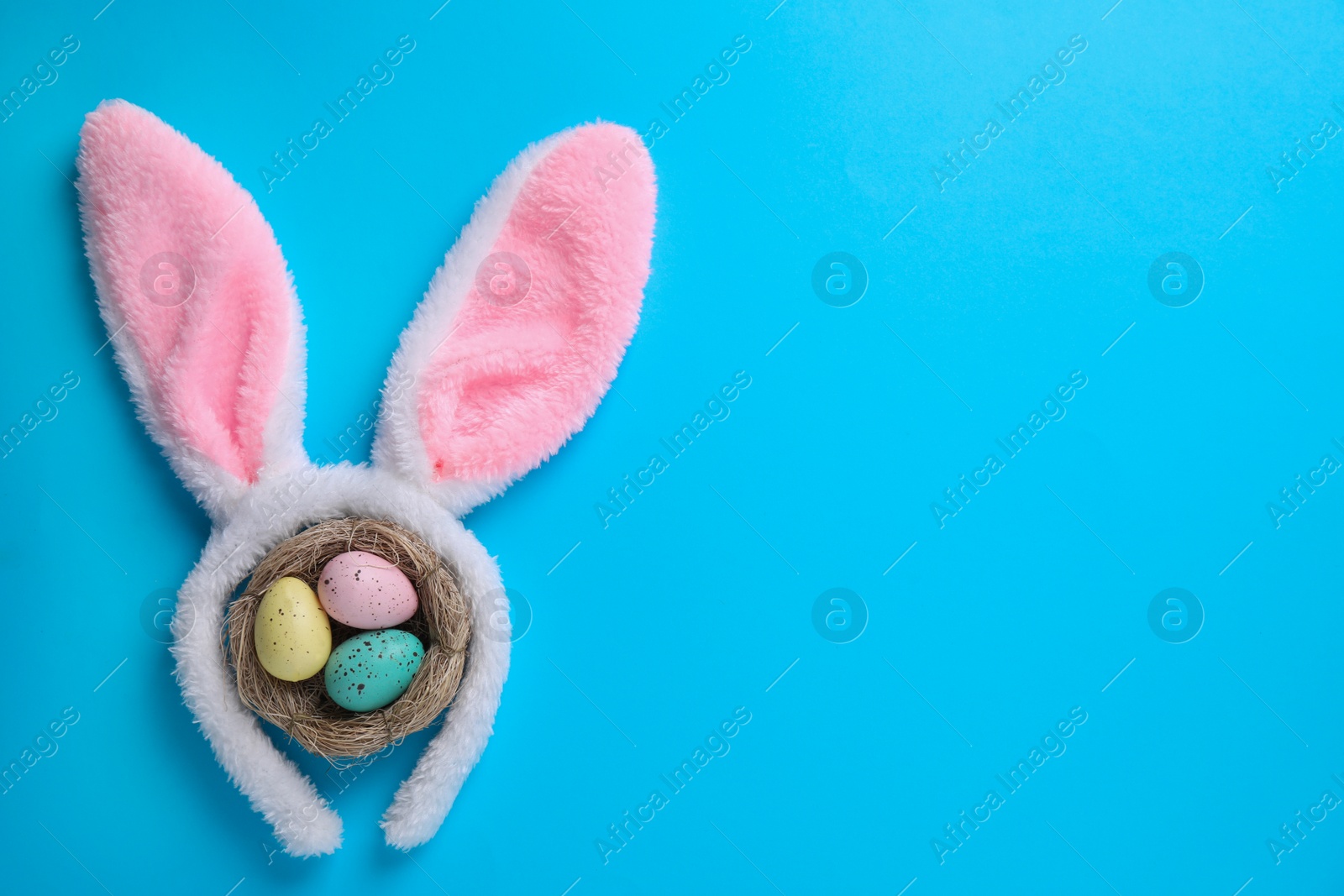 Photo of Headband with Easter bunny ears and dyed eggs in nest on light blue background, flat lay. Space for text