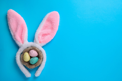 Photo of Headband with Easter bunny ears and dyed eggs in nest on light blue background, flat lay. Space for text