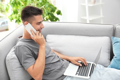 Photo of Man talking by mobile phone while using laptop on sofa at home