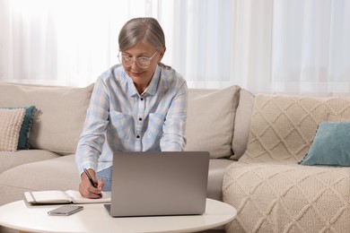 Photo of Beautiful senior woman writing something in notebook while using laptop at home