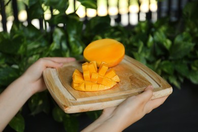 Woman holding wooden board with cut ripe mango outdoors, closeup