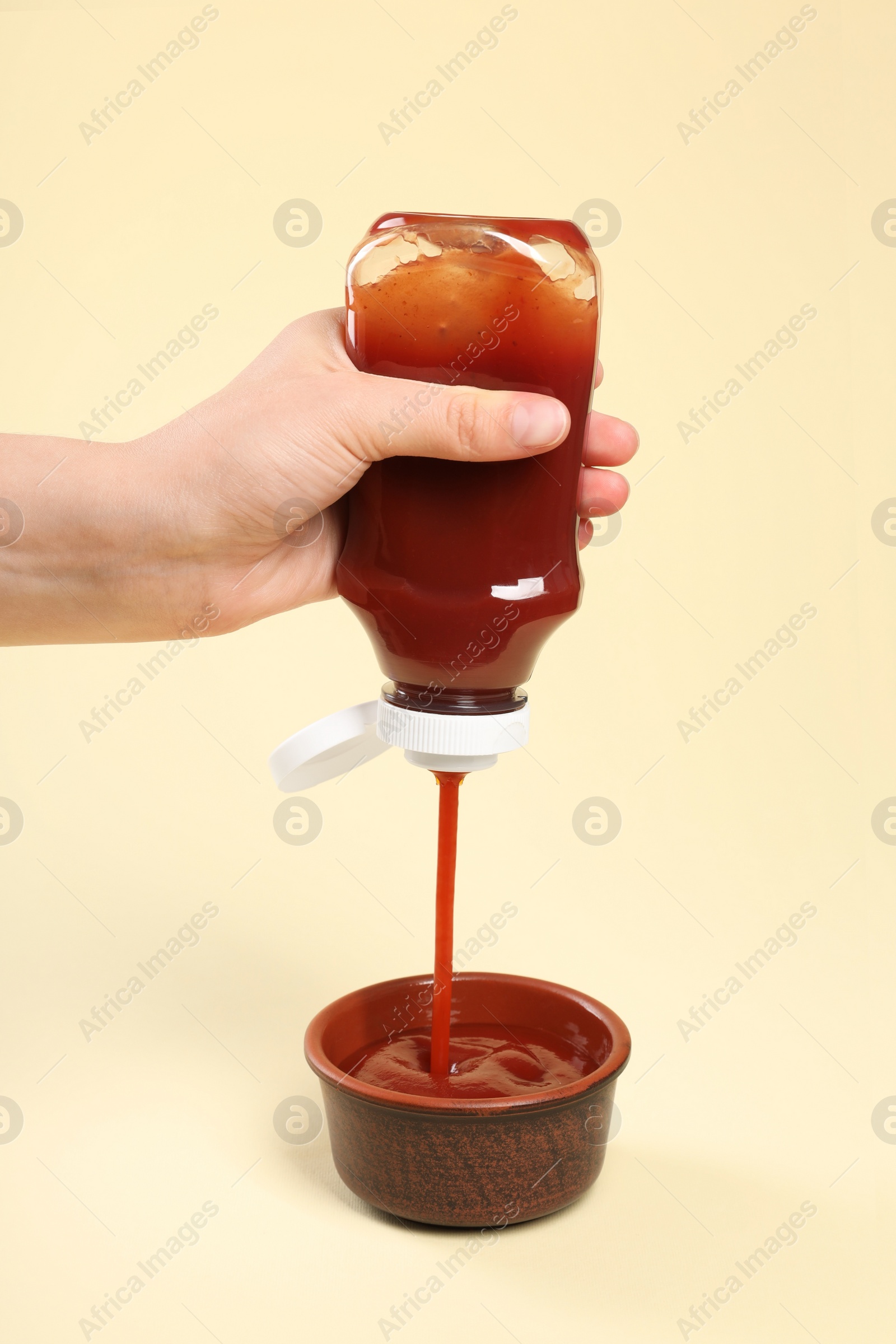 Photo of Woman pouring tasty ketchup from bottle into bowl on beige background, closeup