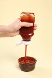 Photo of Woman pouring tasty ketchup from bottle into bowl on beige background, closeup