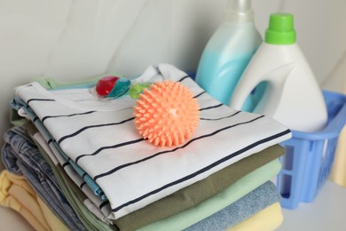 Orange dryer ball and pods on stacked clean clothes near laundry detergents, closeup
