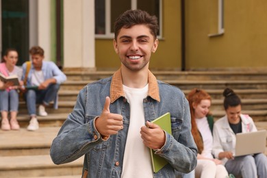 Photo of Students learning together on steps. Happy young man with notebook showing thumbs up outdoors, selective focus