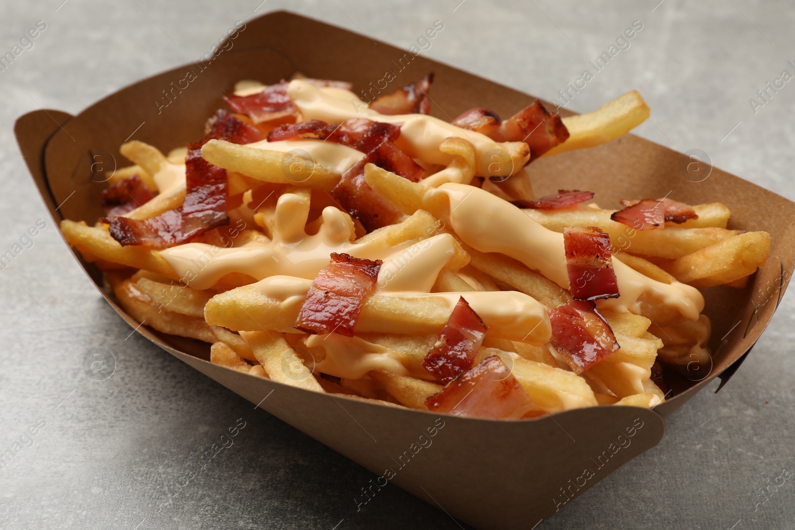 Photo of Tasty potato fries, cheese sauce and bacon in paper container on grey table, closeup