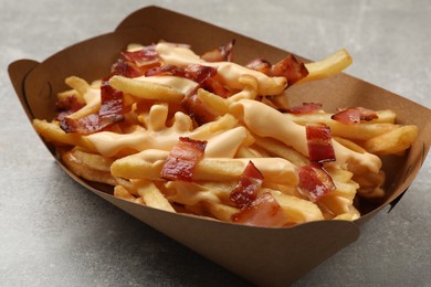 Photo of Tasty potato fries, cheese sauce and bacon in paper container on grey table, closeup