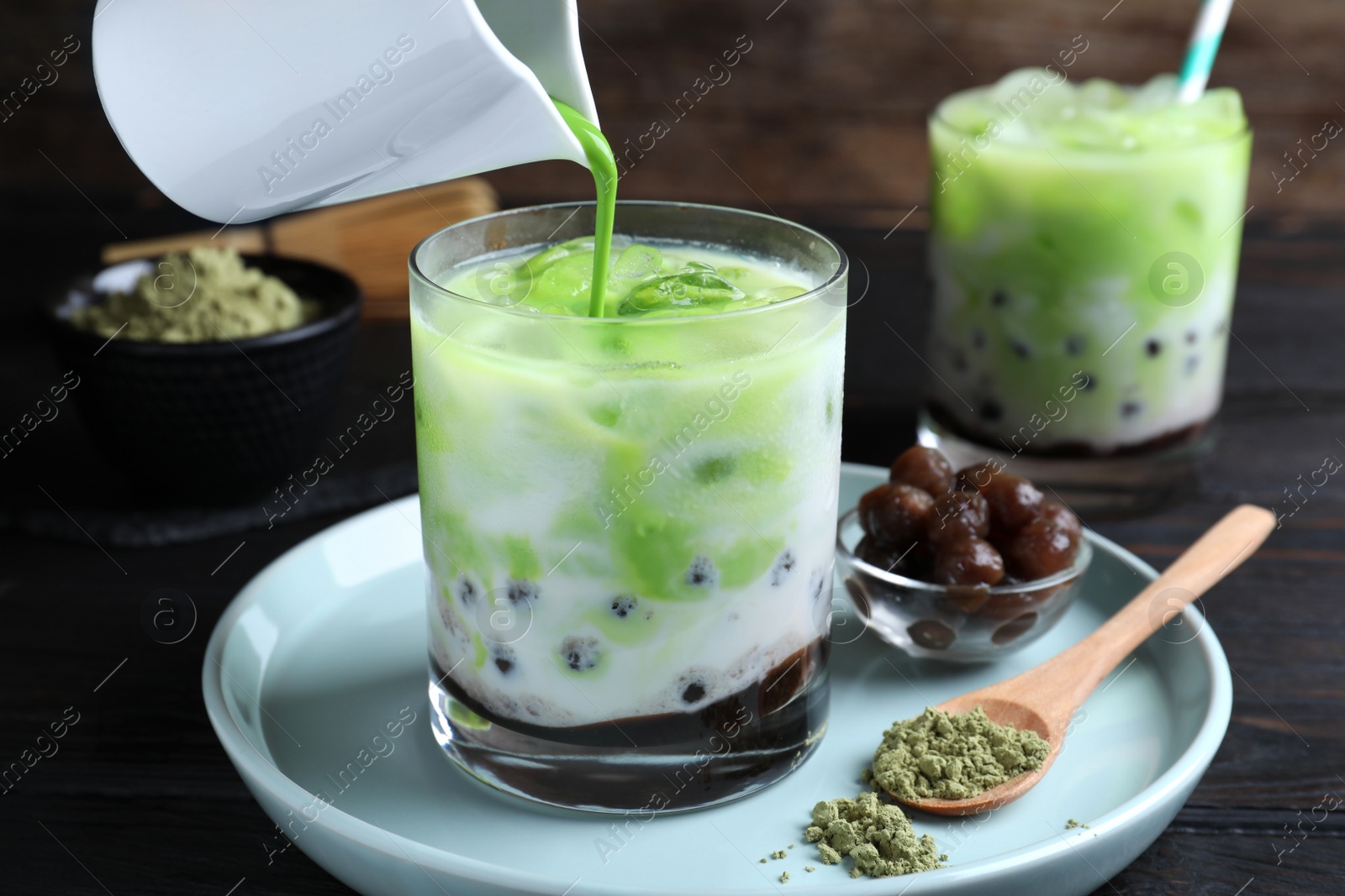 Photo of Pouring green matcha into glass with milk bubble tea at black wooden table, closeup