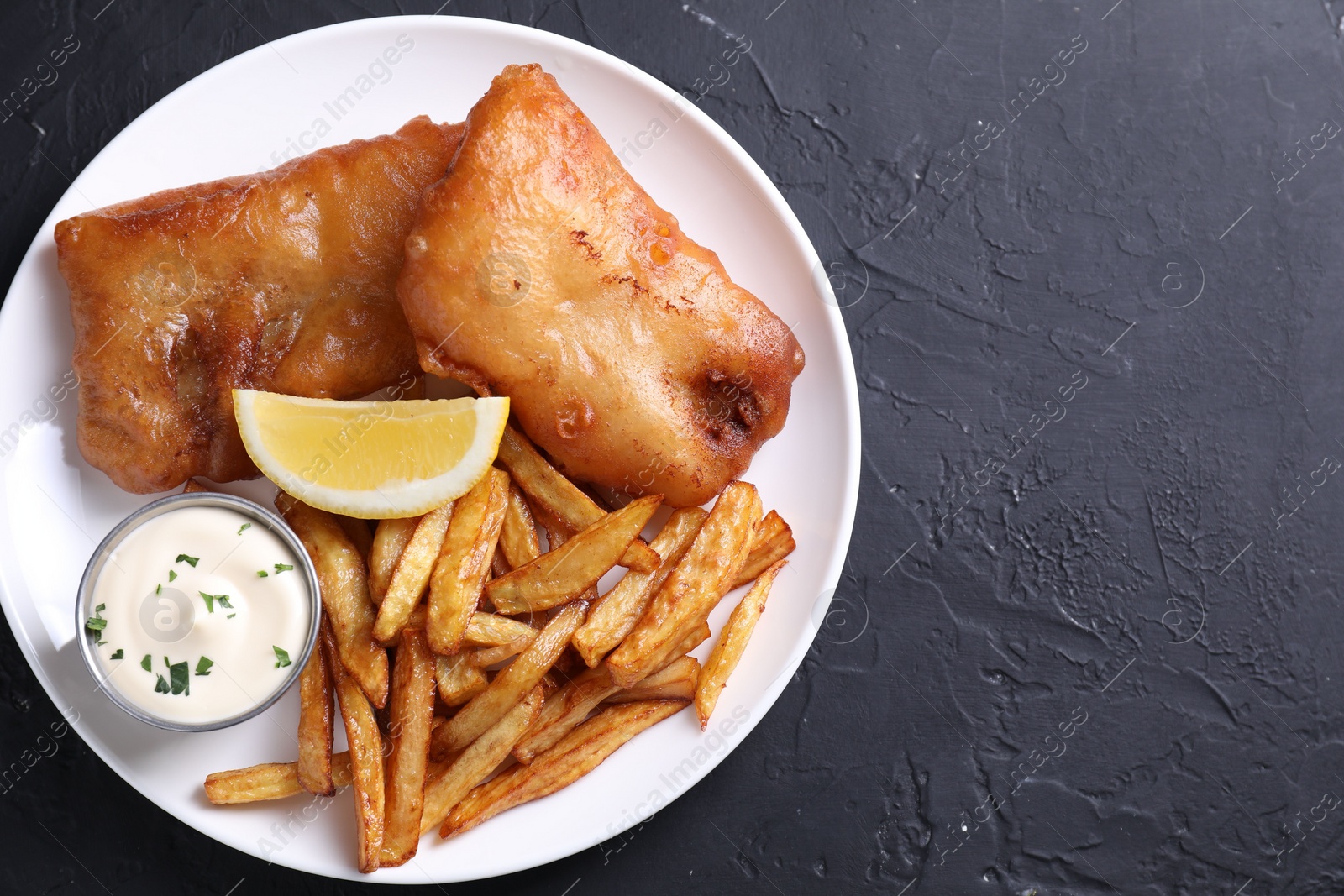 Photo of Tasty fish, chips, lemon and sauce on black table, top view. Space for text