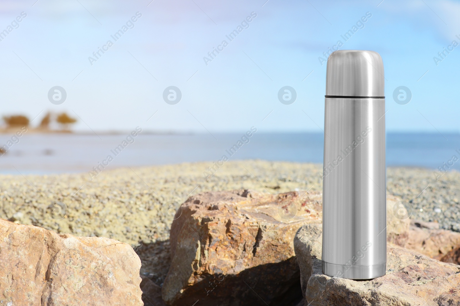 Photo of Metallic thermos with hot drink on stone near sea, space for text