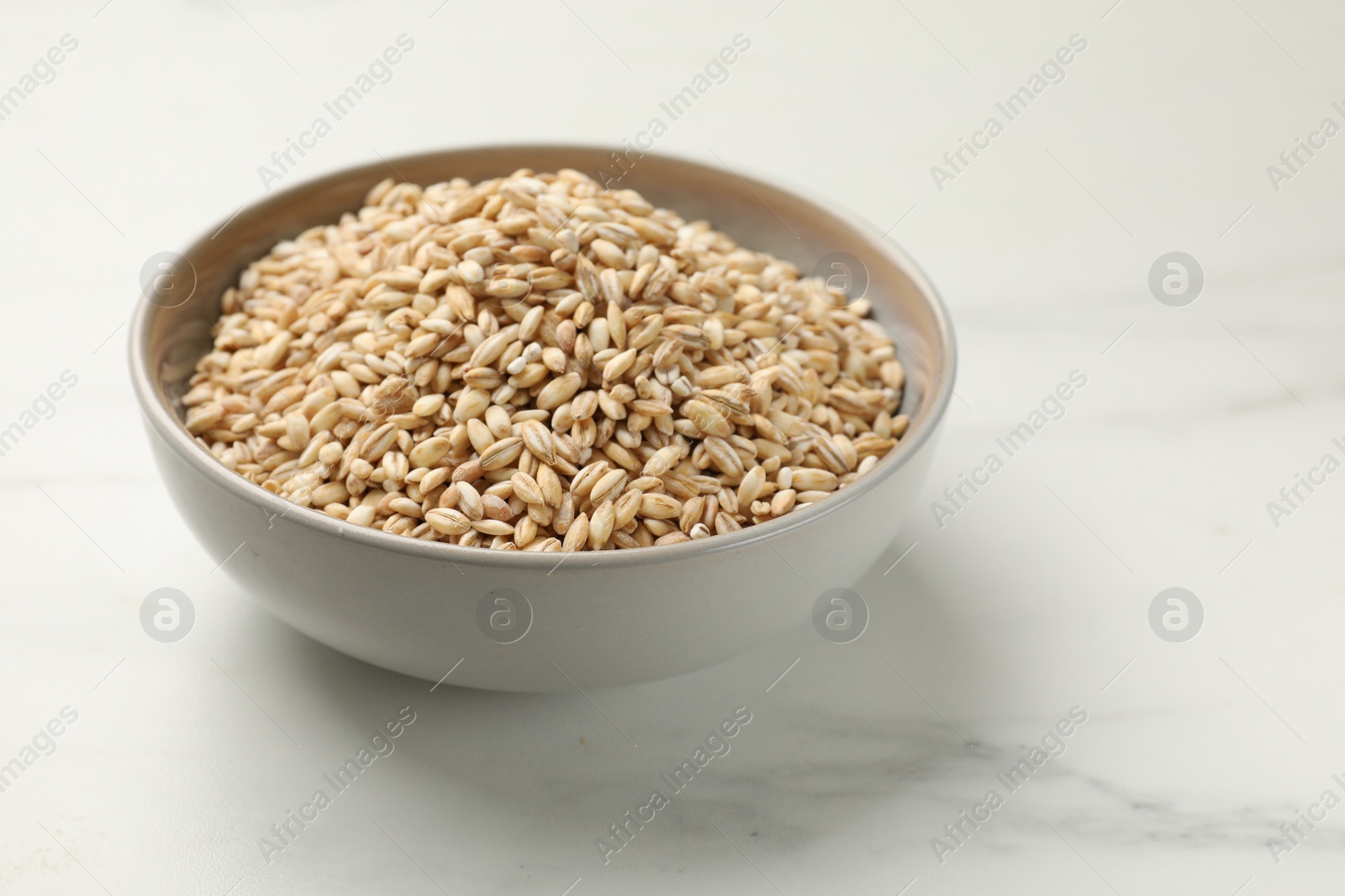 Photo of Dry pearl barley in bowl on white marble table, space for text