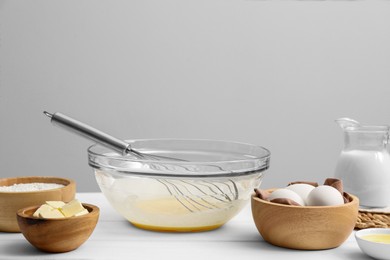 Glass bowl of crepe batter with whisk and ingredients on white wooden table, space for text