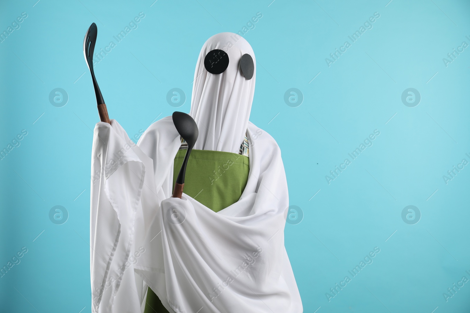 Photo of Woman in ghost costume and apron with kitchen set on light blue background, space for text