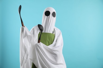 Woman in ghost costume and apron with kitchen set on light blue background, space for text