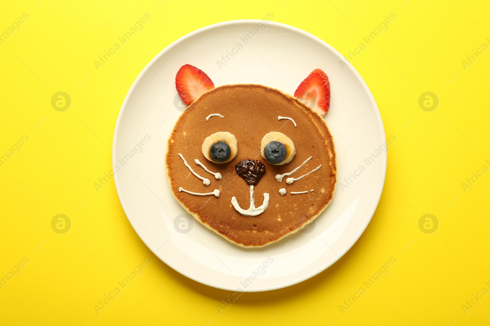 Photo of Creative serving for kids. Plate with cute cat made of pancakes, berries, cream, banana and chocolate paste on yellow background, top view