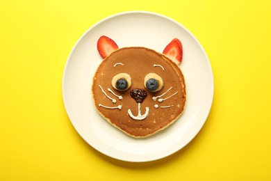 Photo of Creative serving for kids. Plate with cute cat made of pancakes, berries, cream, banana and chocolate paste on yellow background, top view