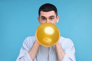 Photo of Young man inflating golden balloon on light blue background