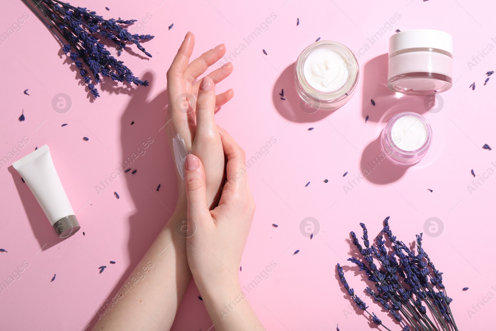 Photo of Woman applying hand cream and lavender flowers on pink background, top view