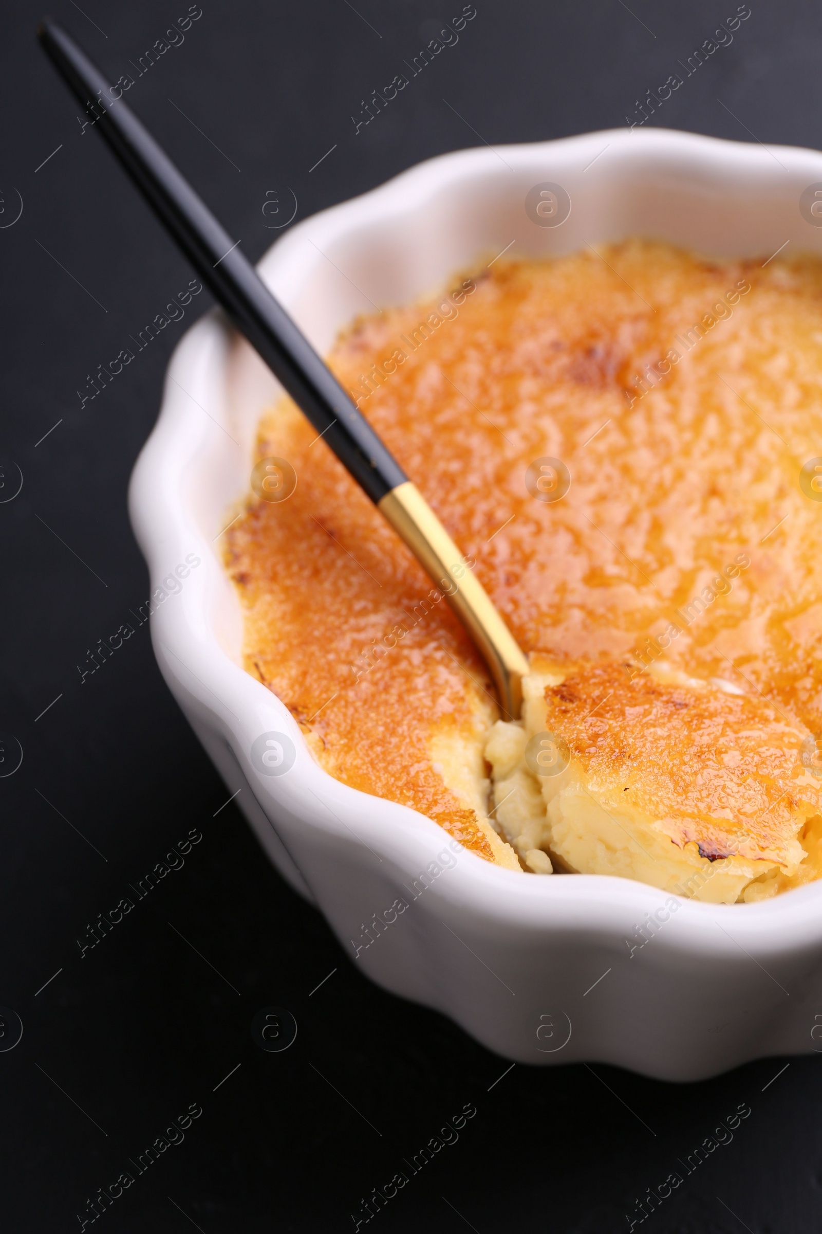 Photo of Delicious creme brulee in bowl and spoon on dark gray table, closeup