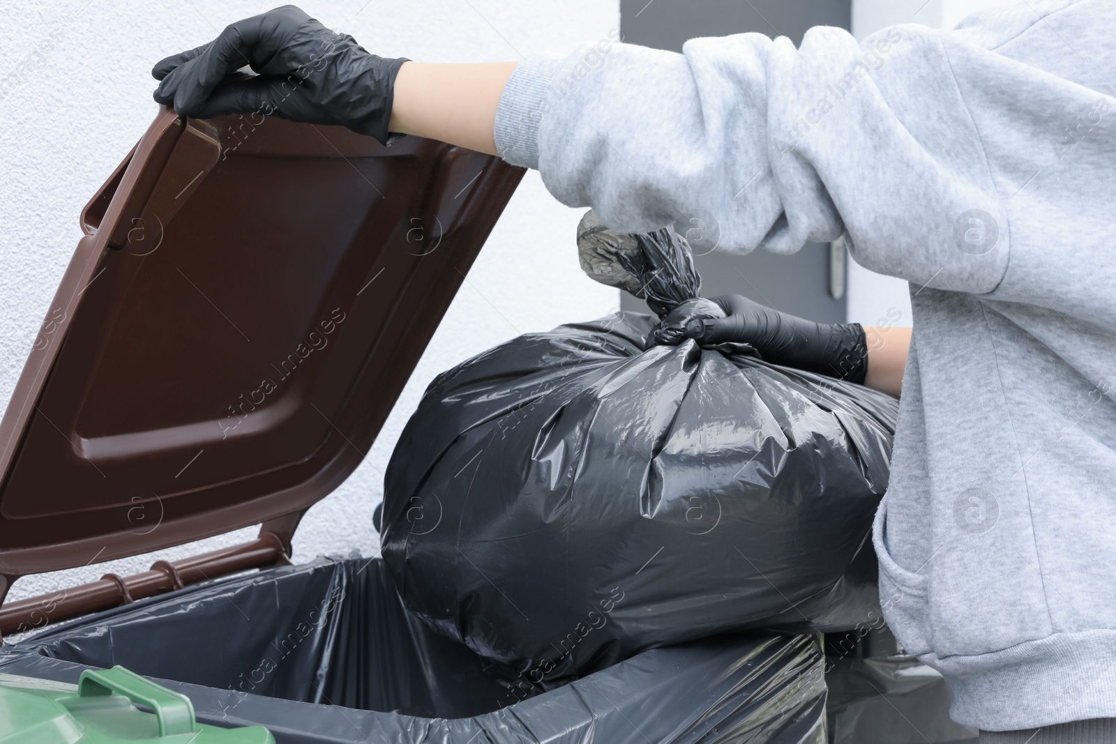 Photo of Woman throwing trash bag full of garbage in bin outdoors, closeup. Recycling concept