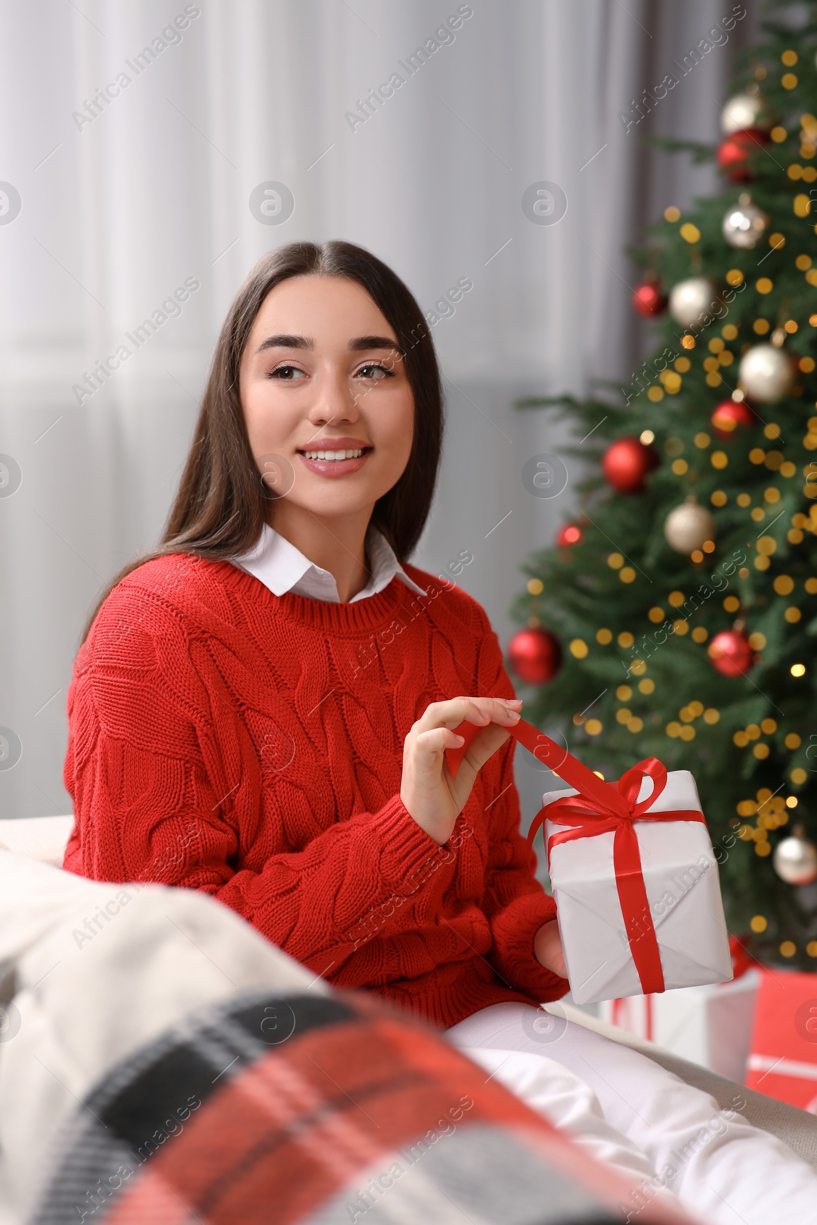 Photo of Smiling woman opening gift near Christmas tree indoors