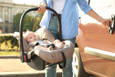 Photo of Mother holding child safety seat with baby near car outdoors
