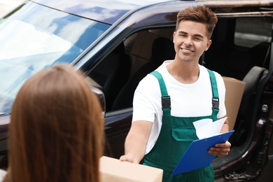 Photo of Young delivery courier with clipboard giving parcel to customer outdoors