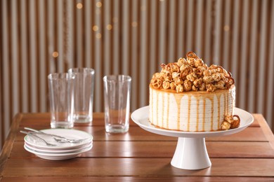 Photo of Caramel drip cake decorated with popcorn and pretzels near tableware on wooden table