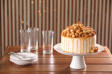 Caramel drip cake decorated with popcorn and pretzels near tableware on wooden table