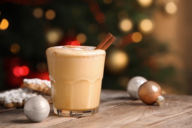 Photo of Tasty eggnog with cinnamon, cookies and baubles on wooden table against blurred festive lights. Space for text
